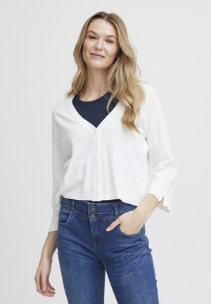 White cropped cardigan with a V-neck, featuring a button front and three-quarter sleeves, paired with dark blue denim jeans.
