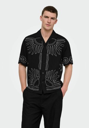 Joven con el pelo corto que lleva una camisa negra de manga corta con botones y patrones abstractos blancos de sol y remolinos, manos en los bolsillos.