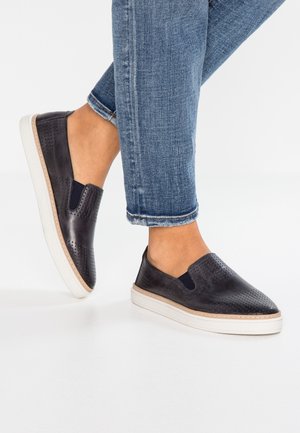 Pieds portant des chaussures noires perforées à enfiler avec semelles blanches, associées à un jean bleu retroussé sur un fond blanc.