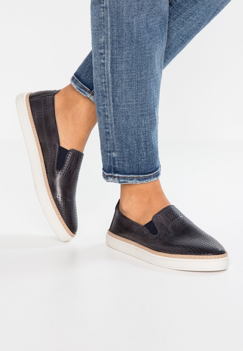 Pieds portant des chaussures noires perforées à enfiler avec semelles blanches, associées à un jean bleu retroussé sur un fond blanc.