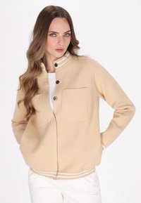 Beige zip-jacka med hög krage, två bröstfickor och vita randiga detaljer. Slät stickad tyg och ribbat avslut.