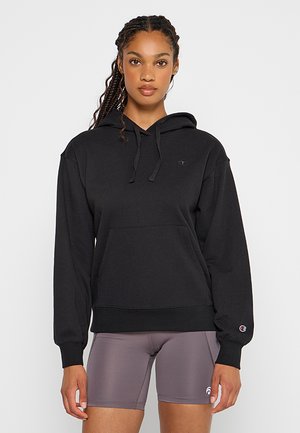 Joven mujer con el cabello trenzado que lleva una sudadera con capucha negra y pantalones cortos ajustados grises, de pie contra un fondo liso y claro.