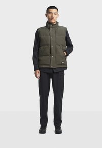 Ärmellose olivgrüne Steppweste mit Druckknöpfen, Fronttaschen, getragen über einem schwarzen Langarmshirt und kombiniert mit schwarzen Cargo-Hosen und Schuhen.