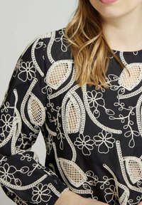 Blouse noire avec broderie florale blanche, détails en coupure et couture texturée. Manches longues avec poignets froncés.