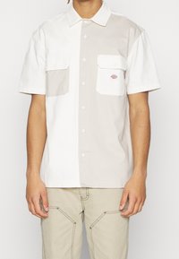 Camicia a maniche corte in cotone bianco e beige chiaro. Presenta due tasche sul petto con pattina e chiusura a bottone, marchio discreto sulla tasca sinistra.