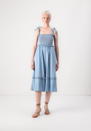 Light blue denim midi dress with smocked bodice, ruffled accents at the waist and hem, and adjustable tied straps. Worn with strappy heels.