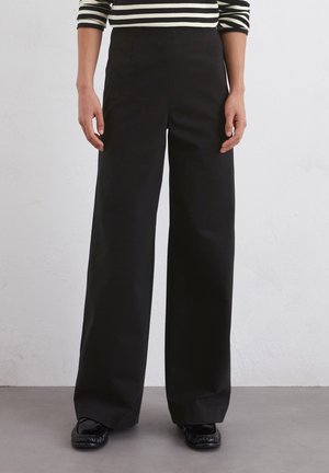 Person trägt schwarze, weit geschnittene Hosen, schwarze Loafers und ein schwarz-weiß gestreiftes Langarmshirt und steht vor einer grauen Wand.