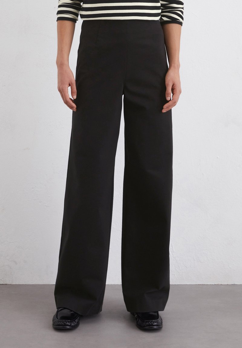 Personne portant un pantalon large noir, des mocassins noirs et un haut à manches longues rayé noir et blanc, debout contre un mur gris.
