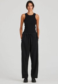 Crna majica bez rukava u kombinaciji s crnim jogger hlačama s elastičnom stražnjom gumicom, koje imaju opušteni kroj i bočne džepove. Model nosi crne cipele.