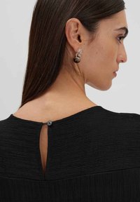 Top nero textured con chiusura a bottoni sul retro, caratterizzato da un dettaglio a chiave. Accompagnato da orecchini rotondi a più strati realizzati con materiali trasparenti e scuri.