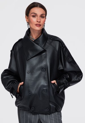 Femme portant une veste en cuir noire oversize à double boutonnage avec grand col, mains dans les poches et pantalon gris rayé.