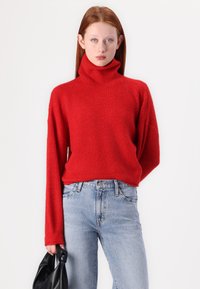 Crvena, teksturirana turtleneck pulover s dugim rukavima u kombinaciji s svijetloplavim hlačama visokog struka. Model drži crnu torbu.