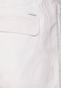Biely textúrovaný materiál s panelom a vreckom. Šitá etiketa s logom "LINDENBERGH". Jednoduchý, funkčný dizajn bez ďalších ozdôb.
