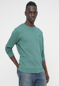 Βεραμάν φούτερ από βαμβάκι με ριμπ μανσέτες και τελείωμα, με μικρό κίτρινο λογότυπο στο στήθος. Συνδυασμένο με μπλε τζιν παντελόνι.