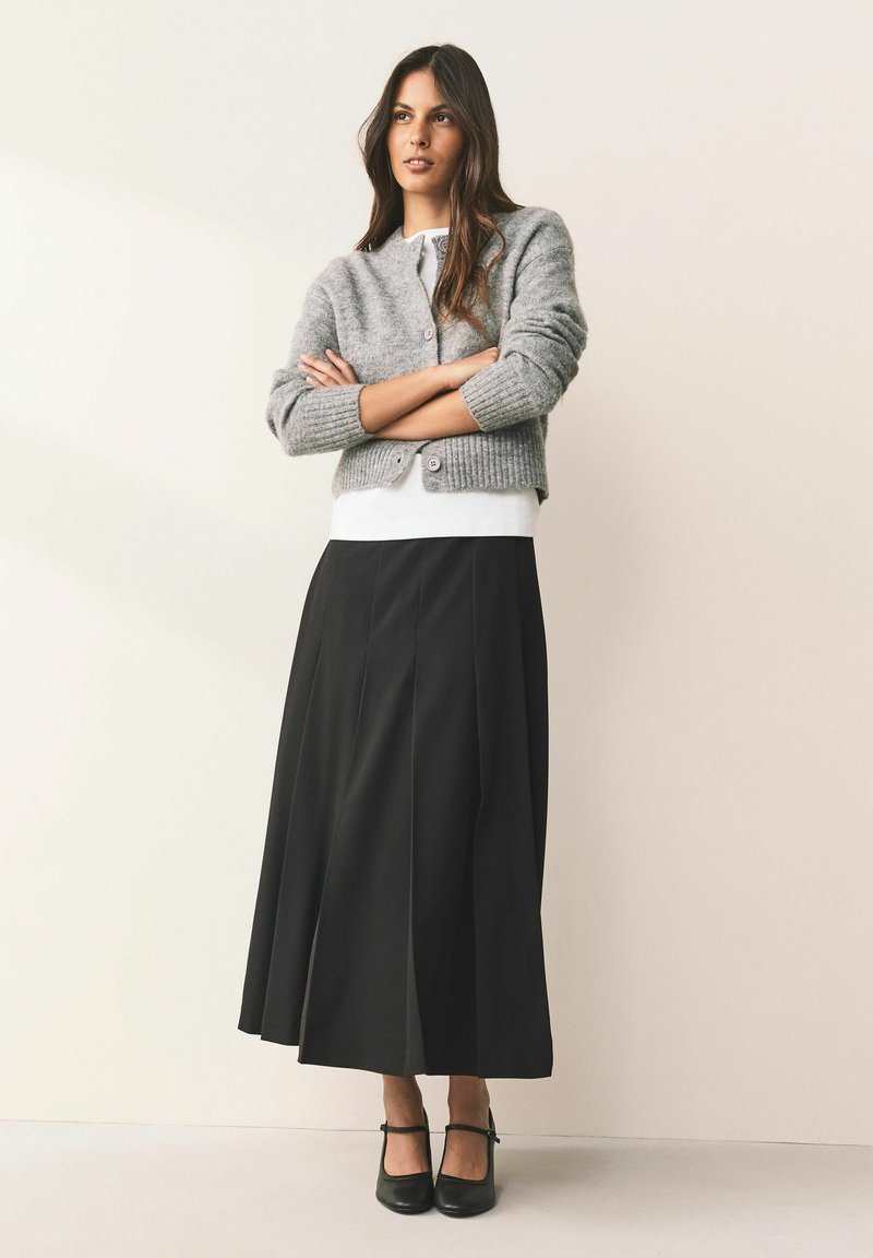 Cardigan gri tricotat cu manșete reiate, top alb și fustă plisată neagră. Pantofi cu toc negri au completat ținuta.