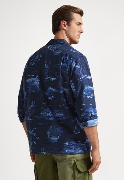 Άνδρας που φοράει μπλε σκούρο πουκάμισο με μακριά μανίκια και γυρισμένα μανίκια, διακοσμημένο με ανοιχτόχρωμες, μπλε εικόνες εξωτερικής περιπέτειας, συνδυασμένο με χακί cargo παντελόνι.