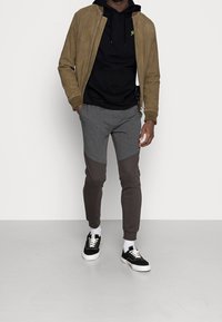 Giacca bomber in pelle scamosciata marrone, felpa con cappuccio nera, pantaloni jogger grigi e grigio scuro, e sneaker nere con suole e lacci bianchi.