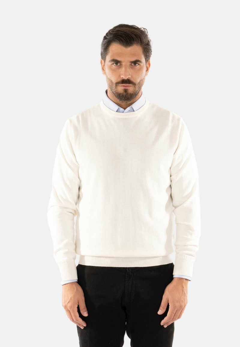 Maglione lavorato a maglia di colore crema, con maniche lunghe, scollatura rotonda e polsini a costine, indossato sopra una camicia azzurra con colletto, abbinato a pantaloni neri.