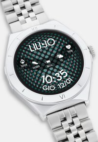 Reloj de acero inoxidable plateado con una esfera negra redonda que presenta un patrón de puntos. La pantalla principal muestra la hora, la fecha y los iconos de aplicaciones y salud.