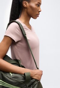 Γυναίκα με ροζ κοντομάνικο πουκάμισο, κρατώντας μια ανοιχτοπράσινη αθλητική τσάντα Puma στον ώμο της, μπροστά σε ένα απλό φόντο.