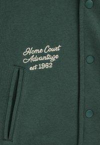 Temno zelena tkanina z belo izvezenim besedilom "Home Court Advantage est. 1962", navpičnim razporkom za žep in zelenimi pritrdilnimi gumbi.