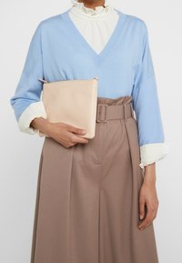 Béžová kožená kabelka držená proti světle modrému oversized svetr s bílým volánkovým límcem, v kombinaci s hněd ými kalhotami s vysokým pasem.