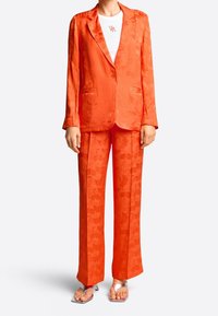 Costume à motifs floraux orange avec une veste ajustée et un pantalon évasé. Tissu lisse, associé à un top blanc et des sandales argentées à lanières.