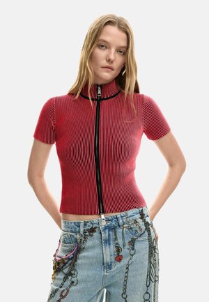 Top roșu cu mâneci scurte, tricotat cu model nervurat, cu guler înalt și fermoar frontal, asortat cu blugi albaștri din denim, decorați cu lanțuri și talismane.