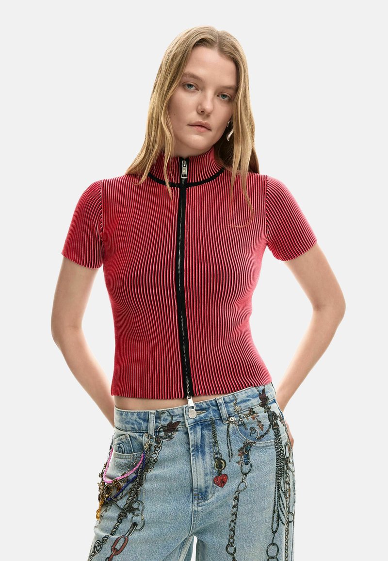 Červený rebrovaný pletený krátky rukáv s vysokým golierom a predným zipsom, v kombinácii s modrými džínsami zdobenými retiazkami a príveskami.
