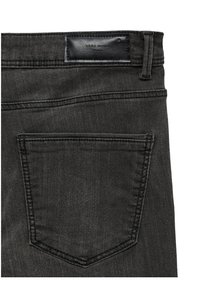 Dos d'un jean en denim noir montrant une poche unique et une étiquette de marque en cuir noir cousue au-dessus de la ceinture.