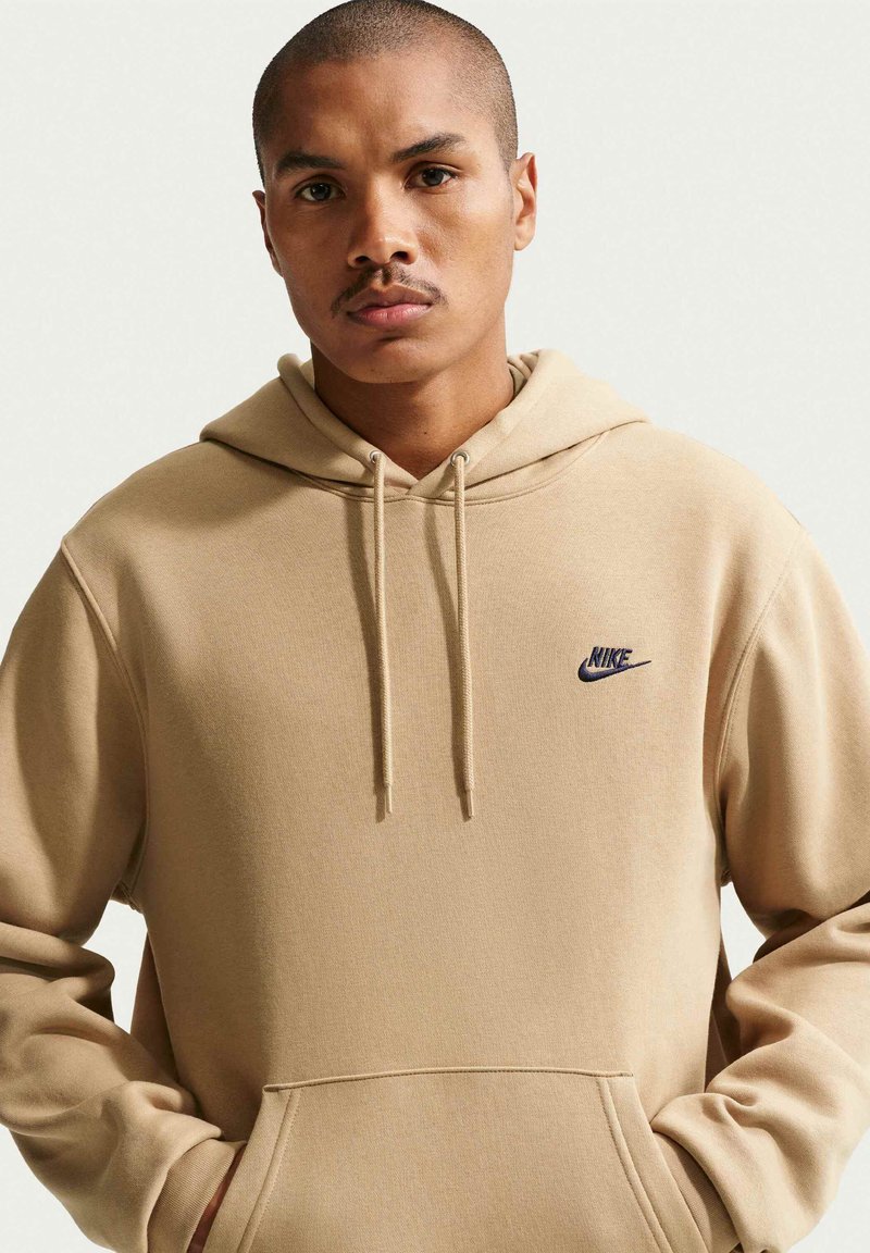 Mann mit kurzem Haar und Schnurrbart, der einen beigen Nike Hoodie trägt und die Hände in der vorderen Tasche hat, vor einem einfarbigen Hintergrund.