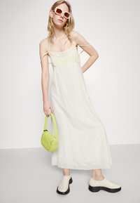 Robe maxi blanche fluide avec un corsage jaune clair, accompagnée de chaussures noires chunky et d'un petit sac à main vert. Modèle portant des lunettes de soleil surdimensionnées.