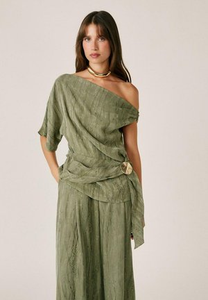 Mujer con vestido verde oliva de un solo hombro con tejido texturizado y detalle de broche dorado, accesorios con collar dorado grueso, posando con las manos en los bolsillos.