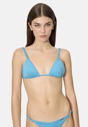 Joven con el pelo castaño liso que lleva un bikini de triángulo azul claro con braguitas a juego, sobre un fondo liso.