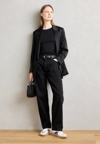 Blazer noir ajusté sur un haut noir, associé à un jean noir taille haute. Les accessoires incluent un petit sac à main noir et des baskets argentées.