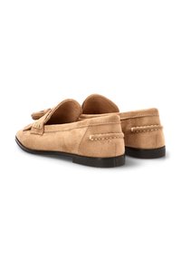 Mocasines de ante en marrón claro, con punta redondeada, suela plana negra, flecos decorativos y detalles de costura en el talón y los laterales.