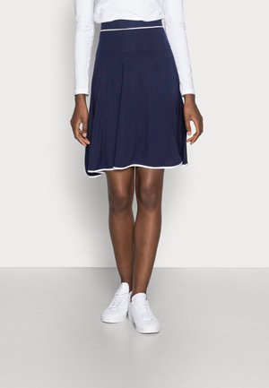 Kobieta ubrana w granatową spódnicę o kroju litery A z białym wykończeniem, białą koszulę z długim rękawem i białe trampki, stojąca na tle jasnego, jednolitego tła.