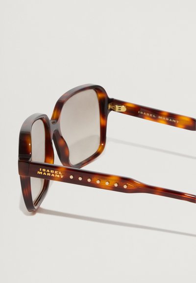 Occhiali da sole tartarugati con montatura quadrata, lenti a gradiente e dettagli dorati sulle astine con piccoli ritagli circolari.