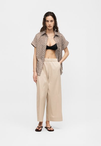 Γυναίκα με μακριά κυματιστά μαλλιά που φοράει κοντομάνικο πουκάμισο με σχέδια ανοιχτό πάνω από μαύρο τοπ, μπεζ παντελόνι με φαρδιά πόδια και μαύρα σανδάλια, στέκεται με το χέρι στην τσέπη.