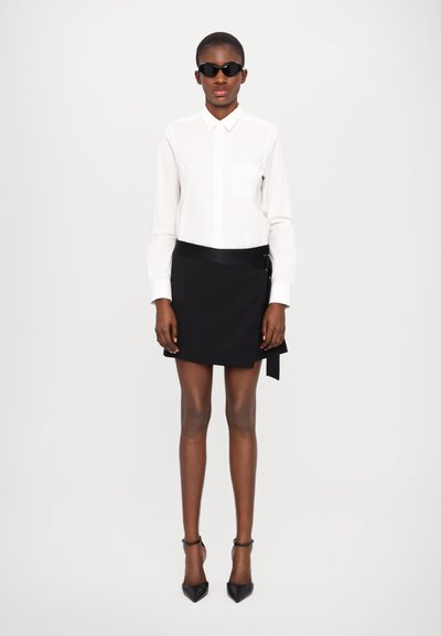 Camicia bianca a maniche lunghe con taschino sul petto, abbinata a una gonna nera a portafoglio. Il modello indossa occhiali da sole neri e tacchi a punta neri.
