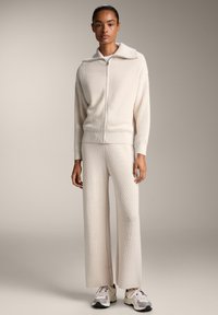 Pletena kremna loungewear kombinacija, ki vsebuje zip-up kardigan z širokim ovratnikom in ohlapnimi, širokima hlačami. Teksturirana površinska podrobnost.