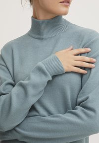 Svetlobluzna rebrasta turtleneck pulover s dolgimi rokavi. Mehak tekstur in oprijet kroj, ki vključuje visok ovratnik in tesne manšete.
