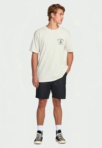 Homem em pé com as mãos nos bolsos, vestindo uma t-shirt branca da Billabong, calções pretos, meias brancas e sapatilhas altas pretas.