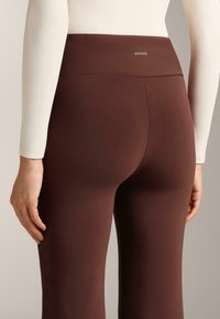 Hnedé legíny s vysokým pásom, hladkej textúry a s logom na páse, nosené s priliehavým krémovým topom s dlhým rukávom.