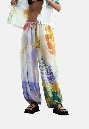 Frau trägt bunte, florale, weite Hosen, schwarze Sandalen und ein schwarzes Bikinioberteil und hebt ein leicht gemustertes Hemd auf.