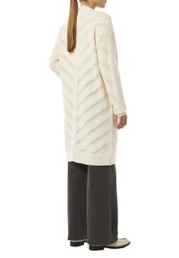 Langer cremefarbener Cardigan mit einem Zickzackmuster, aus strukturiertem Strickstoff gefertigt. Mit geripptem Saum und weiten Ärmeln. Kombiniert mit dunklen weiten Jeans.