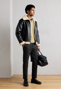 Μαύρο δερμάτινο μπουφάν με εκρού γιακά από shearling, ελιά πουκάμισο, γκρι παντελόνι και μαύρα παπούτσια. Κρατώντας μια μαύρη τσάντα με υφή.