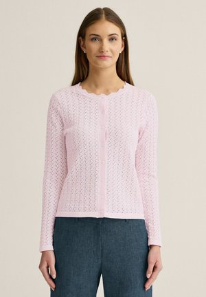Kvinde iført en lyserød tekstureret cardigan med knapper og bølget udskæring samt mørkeblå bukser med høj talje, stående mod en neutral baggrund.