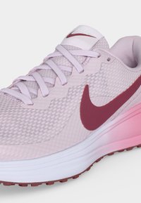 Ανοιχτό ροζ αθλητικό παπούτσι Nike με διχτυωτό πάνω μέρος, επίπεδα κορδόνια, σκούρο κόκκινο λογότυπο swoosh και παχιά λευκή και ροζ σόλα.