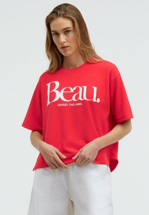 Woman with long brown hair wears a loose red T-shirt with "Beau. SENSES. THE LABEL" in white text and white pants, standing against a plain background.